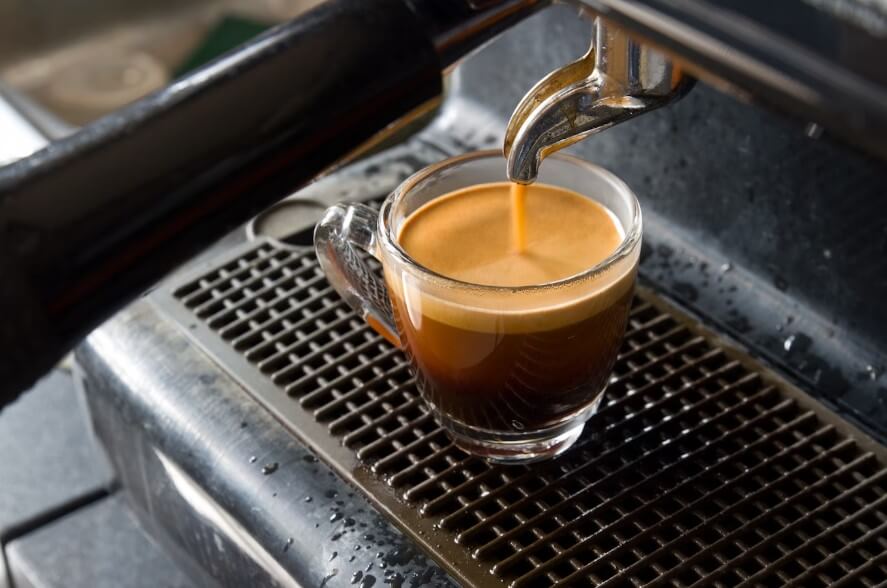 black espresso with crema served in a glass cup next to a coffee machine